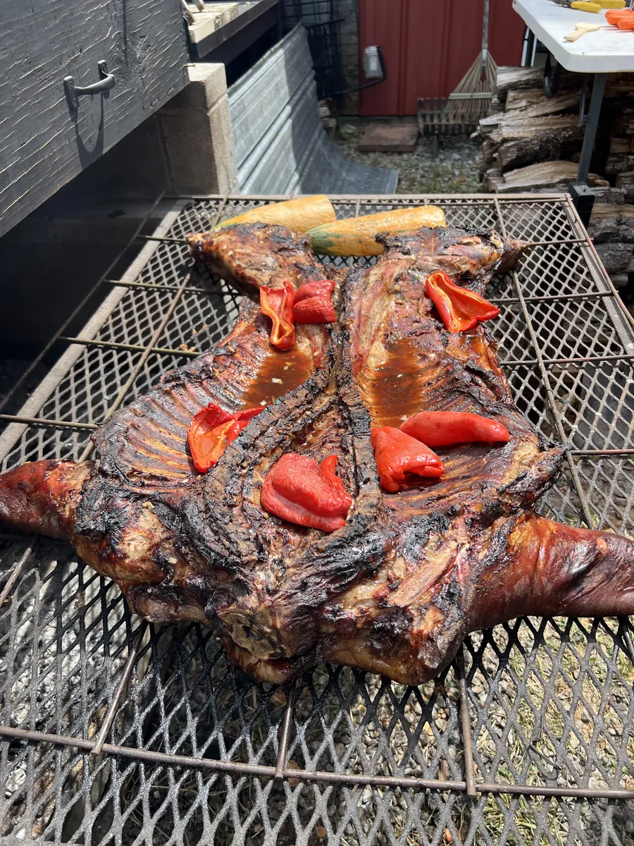 A whole split hog roasted deep brown on a steel grate over the pit, topped with roasted red peppers and yellow squash.